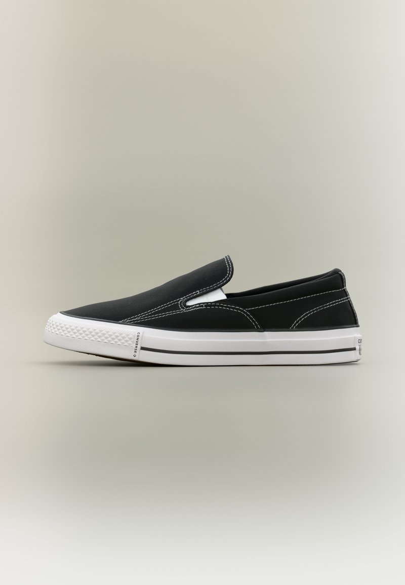 Baskets slip-on noires avec semelle en caoutchouc blanche et surpiqûres blanches contrastantes sur un fond neutre, vue de côté.