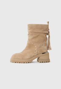 Bottine en suède beige avec des embellissements en clous, un talon épais, une semelle striée et un pompon decoratif à l'arrière.