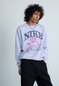 Violeto Nike džemperis ar grafisku attēlu ar spārnu zirgu, ar "Nike" bieza melna teksta un "athletic" zem tā pelēkā krāsā.