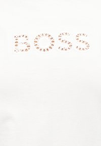 Το όνομα της μάρκας BOSS με κυκλικά μοτίβα κοπής σε κεφαλαία γράμματα σε λευκό ύφασμα.