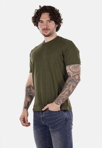 T-shirt in cotone verde oliva con colletto rotondo e maniche corte, indossata da un uomo con tatuaggi, abbinata a jeans in denim blu.