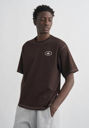 Tânăr purtând un tricou Adidas maro închis și pantaloni de trening gri deschis, stând cu o mână în buzunar, privind în jos spre o parte.