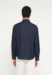 Mężczyzna ubrany w ciemnoniebieską koszulę dżinsową z długim rękawem i jasnobeżowe spodnie, stojący tyłem na tle jednolitego białego tła.