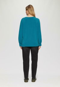Tuerkiser Pullover mit langen Ärmeln und lockerer Passform, kombiniert mit dunklen Hosen und grünen Schuhen, von hinten betrachtet. Glatte Textur, schlichtes Design.