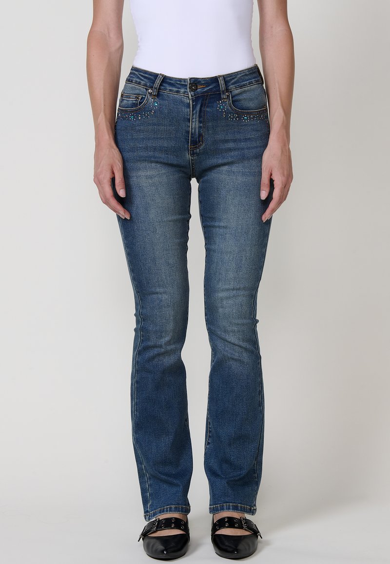 Jeans de mezclilla azules ajustados con una cintura de altura media, que presentan acentos decorativos de strass en los bolsillos frontales y un ligero desgastado en la tela.