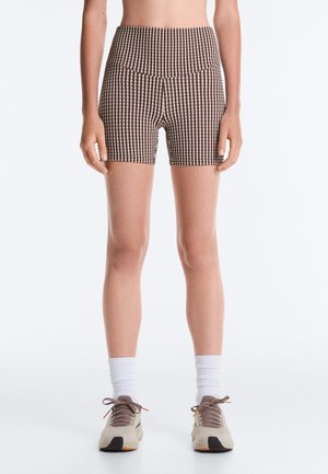 Femme portant un short cycliste à taille haute marron et blanc à carreaux, des chaussettes blanches montantes et des baskets beiges, debout sur un fond blanc uni.