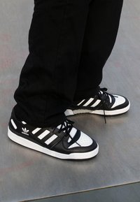 Person wearing black pants and black-and-white Adidas sneakers standing on a smooth gray surface.