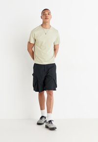 T-shirt vert clair associé à un short noir avec des poches latérales, porté avec des chaussettes blanches et des chaussures noires, se tenant devant un fond blanc.