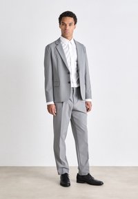 Traje gris hecho de tela suave, con un blazer de solapa muesca y pantalones ajustados. Combinado con una camisa blanca de botones y zapatos negros.
