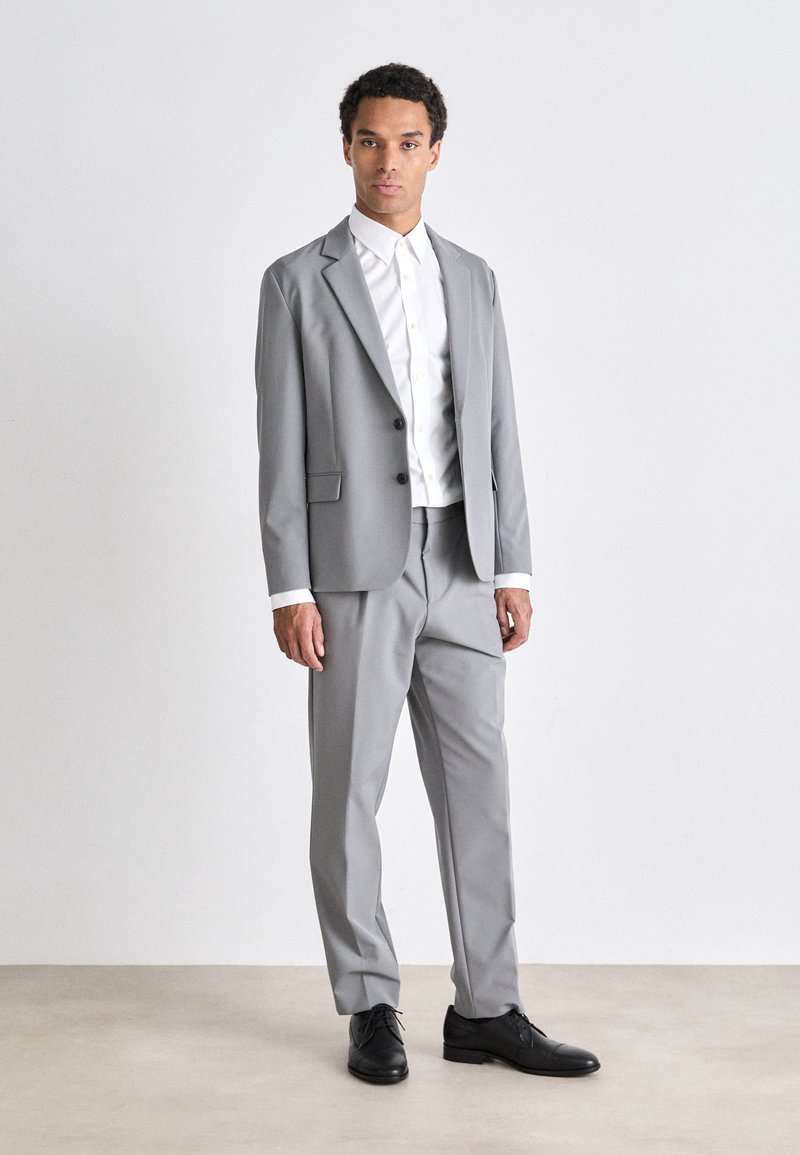 Traje gris hecho de tela suave, con un blazer de solapa muesca y pantalones ajustados. Combinado con una camisa blanca de botones y zapatos negros.