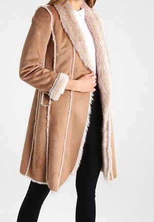 Nainen, joka pukeutunut vaaleanruskeaan mokkatakkiin, jossa on vaalea turkisreuna kauluksessa, hihoissa ja etureunoissa. Yhdistetty mustiin housuihin ja valkoiseen toppiin.