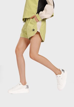 Gambe con pantaloncini verde chiaro e scarpe da ginnastica bianche con dettaglio argento sul tallone, un piede sollevato a metà passo su sfondo neutro.