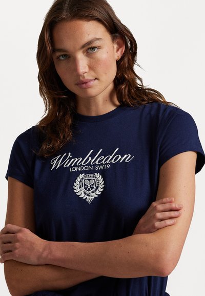Tumši zila kokvilnas t-krekls ar īsām piedurknēm, kuram priekšpusē ir balts izšūts teksts "Wimbledon" un herba dizains. Gluda auduma tekstūra.