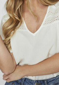 Blouse blanche texturée avec un col en V et des détails décoratifs en œillets le long des épaules, associée à un jean en denim bleu.