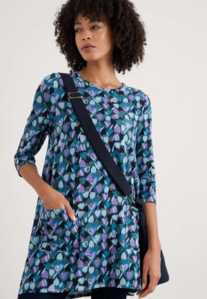 Túnica azul con un patrón floral que presenta tulipanes morados y verdes azules, mangas tres cuartos y bolsillos; el modelo sostiene un bolso negro de hombro.