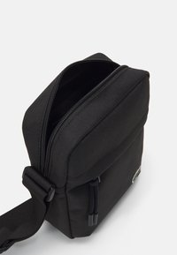 Geantă versatile din material textil negru, tip crossbody, cu închidere cu fermoar, formă rectangulară, curea ajustabilă și branding minim pe partea din față.
