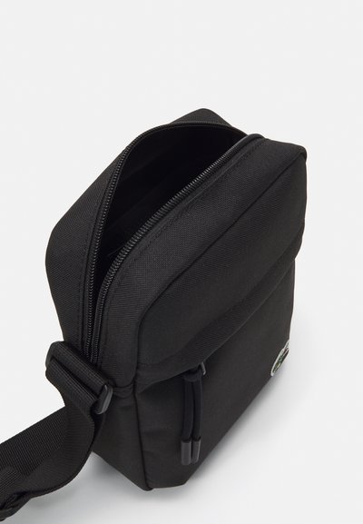 Sac en bandoulière en tissu noir avec une fermeture à zip, de forme rectangulaire, une sangle ajustable et un branding minimal à l'avant.