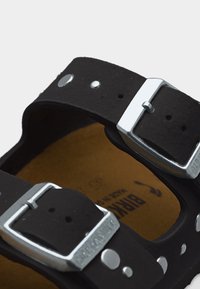 Κοντινό πλάνο μαύρου σανδαλιού με δύο ρυθμιζόμενες ασημένιες αγκράφες και ορατό καφέ εσωτερικό πάτο με τη μάρκα "Birkenstock".