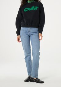 Persoon die een zwarte sweatshirt draagt met groene "Chapot"-tekst, lichtblauwe spijkerbroek en zwarte loafers met studs, staand tegen een witte achtergrond.