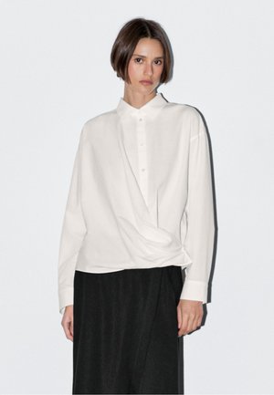 Witte blouse met lange mouwen en knoopsluiting, met een gedrapeerd ontwerp aan de voorkant. Gladde textuur en losse pasvorm, gecombineerd met een zwarte rok.