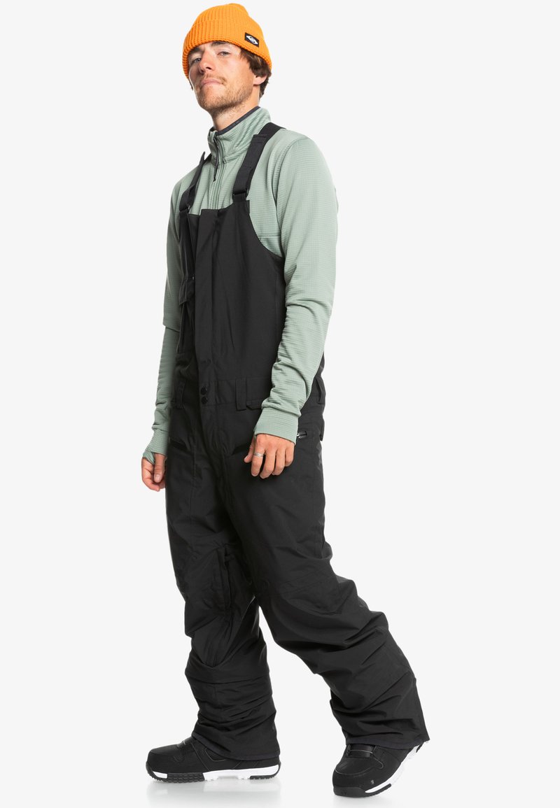 Pantalones de nieve negros con tirantes ajustables, bolsillos laterales y un corte holgado, combinados con una camisa de manga larga verde claro y un gorro naranja.