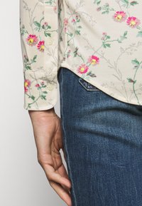 Cremefarbendes Hemd mit Blumenmuster, versehen mit kleinen rosa und grünen Blumen, mit langen Ärmeln und Knopfmanschetten, kombiniert mit blauen strukturierten Jeans.
