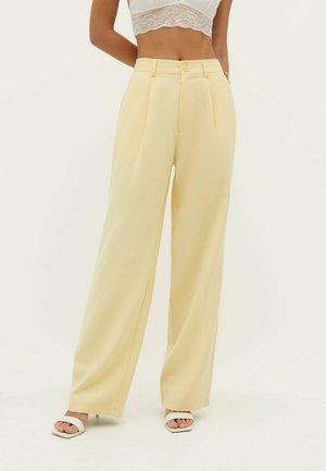 Kvinde iført lysegule bukser med høj talje og vide ben, kombineret med hvide sandaler med hæl og en hvid blonde crop top.