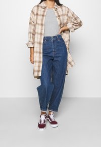 Persona che indossa una lunga camicia beige a scacchi sopra una maglietta grigia, jeans blu a vita alta con risvolti alle caviglie e sneakers bordeaux con lacci bianchi.