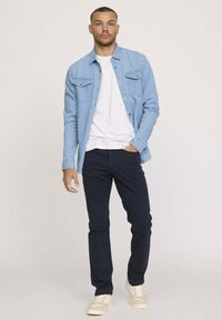 Chemise en denim bleu clair portée sur un t-shirt blanc, associée à un pantalon bleu marine foncé et des baskets crème ; possède des poches boutonnées et une coupe décontractée.