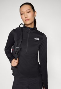 Frau trägt eine schwarze North Face Zip-up-Jacke und schwarze Hose, hält einen schwarzen Rucksackriemen über einer Schulter und schaut nach vorne.