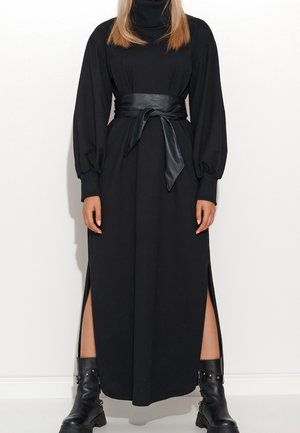 Mujer con vestido negro de manga larga con aberturas laterales, un cinturón ancho negro atado a la cintura y botas negras hasta el tobillo con hebillas.