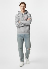 Felpa grigia con un design a cuore sul petto, abbinata a jeans azzurri strappati e sneakers bianche. Il modello è in piedi con le braccia incrociate.