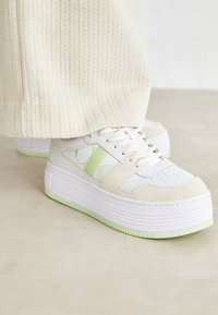 Valkoiset ja beige-kappaleet/platform-kengät, joissa on vihreä korostuslogo. Ominaisuuksiin kuuluvat sileä nahka- ja mokkapinta, rei'itetty varvasosa ja pehmustettu pohja.
