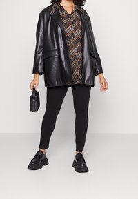 Blazer oversize in ecopelle nera sopra una camicia con motivo, abbinato a leggings neri e scarpe nere chunky, mentre tiene una piccola borsa nera.