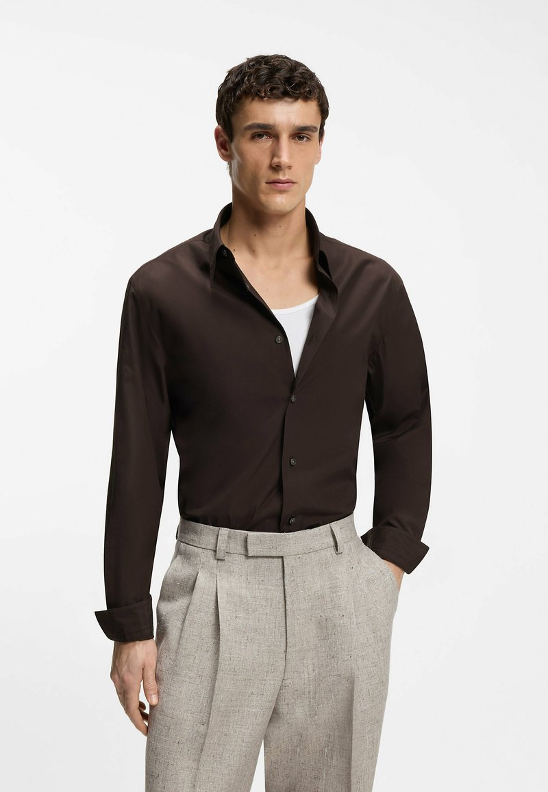 Homme portant une chemise marron foncé boutonnée sur un sous-vêtement blanc, avec un pantalon plissé gris clair, main dans la poche, arrière-plan neutre.