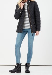 Quilted black jacket with gold hardware, zip and snap closures, paired with a striped top and light blue skinny jeans, complemented by black ankle boots.