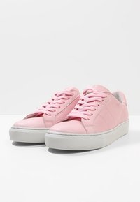 Růžové kožené tenisky s bílými gumovými podešvemi, kulatou špičkou a texturovanými tkaničkami. Hladký povrch s jemnými švy.