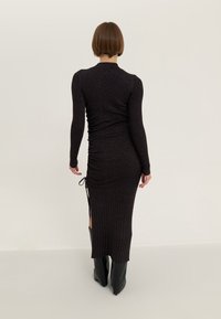 Robe noire à manches longues côtelée avec un design froncé, une fente latérale et un détail de lien ajustable, associée à des bottes noires montant jusqu'aux genoux.