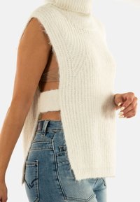 Donna che indossa un maglione estivo bianco senza maniche, con aperture laterali, jeans blu e un reggiseno beige visibile sotto.