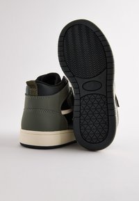 Chaussures montantes vert foncé avec des accents noirs, semelle extérieure en caoutchouc texturé et semelle intermédiaire beige clair. Présentent un design cousu et une languette au talon.