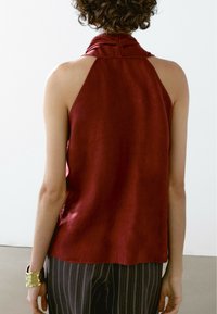 Personne portant un haut sans manches rouge à col licou et un pantalon à rayures fines, avec des cheveux courts et bouclés, debout à l'intérieur contre un mur blanc.