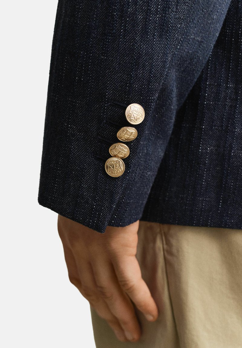 Nahaufnahme einer Hand neben einem dunkelblauen Blazerärmel mit vier goldgeprägten Knöpfen, kombiniert mit beigen Hosen.