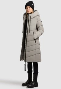 Abrigo de plumas gris claro con capucha, que presenta un patrón en forma de chevrón, bolsillos con cremallera y detalles en negro, combinado con un gorro negro y botas.