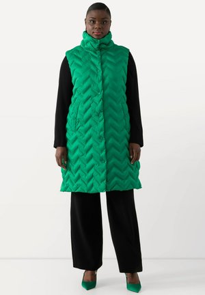 Groene puffer vest met een hoge kraag en een chevron-gequilt patroon, voorzien van knopen en zijzakken, gedragen over zwarte kleding.