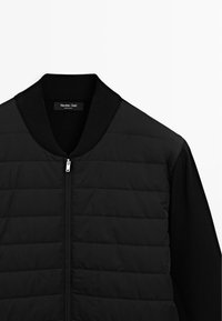 Schwarze Zip-Up-Jacke mit geripptem Kragen. Der Oberkörper weist ein gestepptes Muster auf; Ärmel und Rücken bestehen aus einem glatten, strukturierten Stoff.