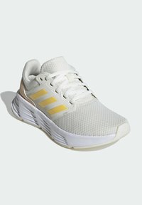 Scarpa sportiva bianca con una tomaia in mesh testurizzata, dettagli dorati e tre strisce orizzontali. Presenta una suola imbottita e un design con lacci.