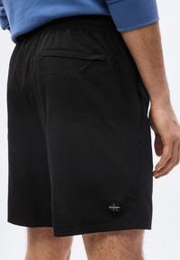 Homme portant un short noir Calvin Klein avec poche zippée au dos et taille élastique, vu de dos au niveau des cuisses.