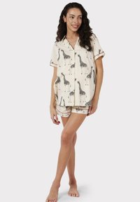 Conjunto de pijama de manga corta en crema con patrones de jirafas negras, que incluye una camisa con botones y pantalones cortos a juego con adornos decorativos.