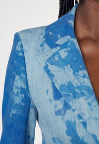 Personne à la peau foncée portant un blazer en denim bleu avec un motif décoloré et tacheté et un col à revers cranté.