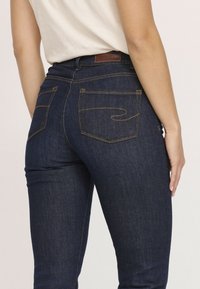 Jean en denim foncé avec une coupe ajustée, poches standards avec des coutures jaunes, et une étiquette en cuir marron au niveau de la taille.